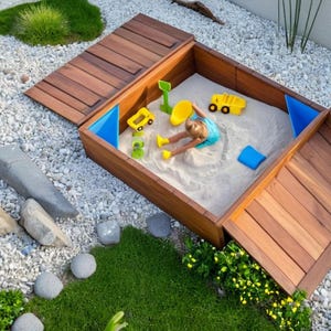 May include: A wooden sandbox with a hinged lid, open to reveal a child playing with toy trucks and other toys in the sand. The sandbox is surrounded by white gravel and green landscaping.