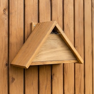 May include: A wooden bat house with a triangular roof is mounted on a vertical wooden surface. The bat house is made of light brown wood and has a small opening. The background is a wooden fence and green foliage.