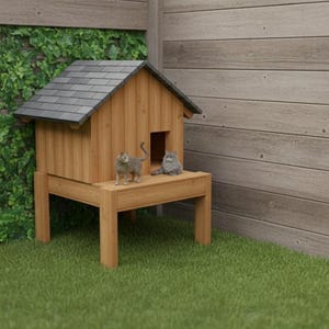 May include: A wooden cat house with a grey tiled roof and a raised platform. Two cats are on the platform. The house is on a green lawn, with a wall of green foliage and a wooden wall in the background.