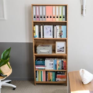 May include: A wooden bookshelf with five shelves. The top shelf has colourful file folders. The second shelf has books and a framed photograph. The third shelf has a printer. The fourth shelf has books. The bottom shelf has books.