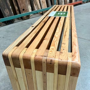 以下が含まれることがあります： リサイクルされた木材の板で作られた木製ベンチ。ベンチはスラットデザインと丸みを帯びたエッジを持っています。木材は薄い茶色です。