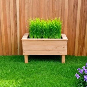 May include: A wooden planter box filled with green grass sits on a bed of artificial green turf. The planter is in front of a wooden fence. A black metal chair and table are on the left, and purple flowers are on the right.