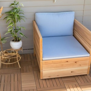 May include: A wooden chair with a light blue cushion sits on a wooden deck. The chair has a storage compartment underneath the cushion.