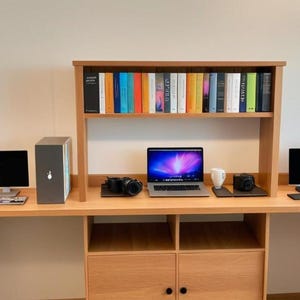 Puede incluir: Un escritorio de madera clara con una estantería incorporada, que contiene una colección de libros coloridos. El escritorio cuenta con un portátil, dos monitores, una taza de café y dos cámaras. Un gran armario de madera está a la izquierda.