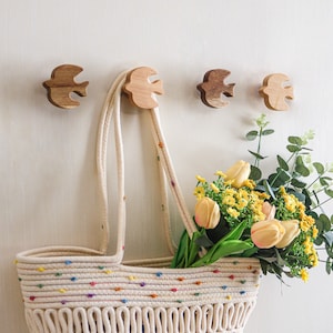 Op de afbeelding: Een witte touwtas met kleurrijke accenten hangt aan een muur met houten vogelhaken. Een boeket gele tulpen met groene bladeren zit in de tas.