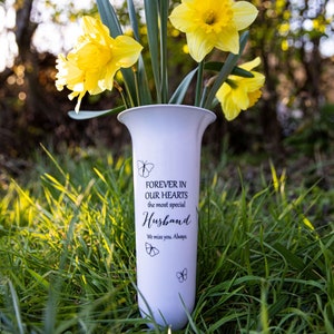 May include: White ceramic vase with a message that reads "Forever in our hearts the most special Husband. We miss you. Always."  The vase is decorated with butterfly silhouettes and is surrounded by green grass and yellow daffodils.