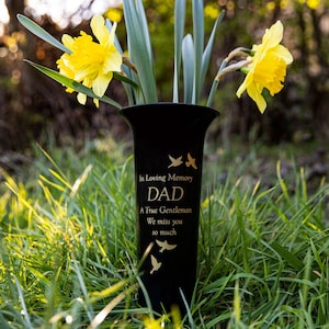 Könnte beinhalten: Schwarze Keramikvase mit goldener Aufschrift "In Loving Memory Dad A True Gentleman We miss you so much". Die Vase ist mit goldenen Vögeln verziert und von grünem Gras und gelben Narzissen umgeben.