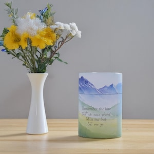 Könnte beinhalten: Eine weiße Vase hält einen Strauß gelber, weißer und blauer Blumen. Daneben befindet sich ein zylindrischer Behälter mit einer Aquarell-Berglandschaft und dem Text: "Remember the love that we once shared. Miss me but Let me go."