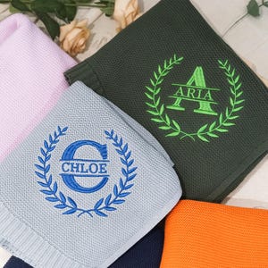 May include: Three knitted baby blankets with personalized embroidery. The blankets are in light blue, light pink, and dark green. The embroidery features a wreath with a letter and a name, "Chloe", "Aria", and a third name that is not visible.