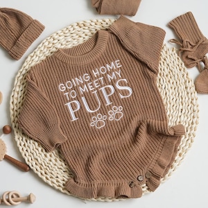 May include: A brown knitted baby outfit set, including a romper with the text "GOING HOME TO MEET MY PUPS", a beanie, and booties. The romper has a ruffled hem and button details. Wooden toys are also visible.