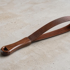 May include: A dark brown wooden handle attached to a textured brown leather strap. The handle features a smooth, polished finish with a circular opening at the end, while the leather strap is looped and secured to the wooden component.
