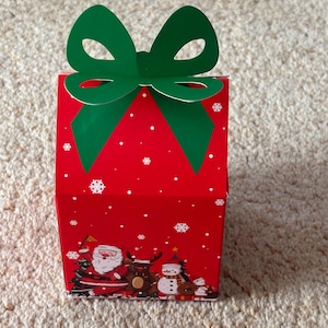 May include: A red Christmas gift box with a green bow. The box is decorated with snowflakes and festive illustrations of Santa Claus, a reindeer, and a snowman.