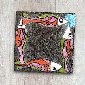 Könnte beinhalten: Eine schwarze quadratische Fliese mit einem farbenfrohen Design aus vier Fischen. Die Fische sind weiß umrandet und haben orange, rosa, grüne und blaue Details. Die Fliese ist ein dekoratives Stück für Haus oder Garten.