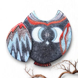 May include: A ceramic owl figurine with a textured, crackled finish. The owl features a dark gray body, white eyes, and accents of red, blue, and brown. The owl's design includes a stylized wing and a decorative base.