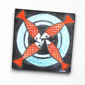 Könnte beinhalten: Eine schwarze quadratische Fliese mit einem blauen und weißen Kreisdesign in der Mitte. Vier orangefarbene Fische mit weißen Augen und Schuppen sind um den Kreis angeordnet, wobei ihre Schwänze nach außen zeigen.