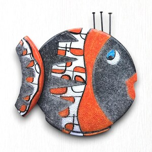 May include: A ceramic fish wall hanging with a black, white, and orange geometric design. The fish has a blue eye and a small black mouth.