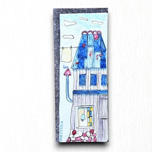 May include: A hand-painted ceramic tile depicting a blue house with a red roof and a chimney. The house has a window with a yellow frame and a bell hanging inside. There are clothes hanging on a line outside the house. The tile has a white background with blue clouds.