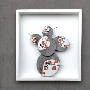 May include: A white framed wall art piece with five round, gray, ceramic discs. Each disc has a geometric design with black lines and orange squares. The discs are attached to the white background with small metal pins.