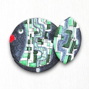 Op de afbeelding: Een zwarte en witte keramische vis met groene en zwarte abstracte patronen. De vis heeft een rode mond en een zwart oog.