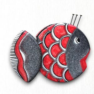 May include: A red and black fish-shaped wall decoration with a white background. The fish has a red body with white scales and a black head and tail.