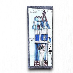 May include: A hand-painted ceramic tile featuring a blue and white house with a red roof and a windmill. The tile has a white background and a black border. The text "ETIVA LAVA ROCK" is written on the tile.