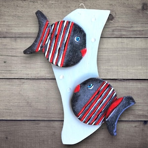 May include: A decorative wall hanging featuring two stylized fish. The fish are black with red and white stripes, red lips, and blue eyes. They are mounted on a light blue, curved backing, creating a nautical theme.
