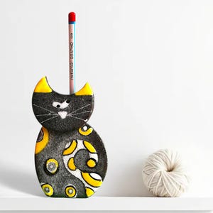 May include: Black and yellow ceramic cat pencil holder with a pencil sticking out of the top.