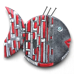 May include: A ceramic fish wall hanging with a black and white geometric pattern on one side and a red and white geometric pattern on the other side. The fish has a blue eye and a red mouth.