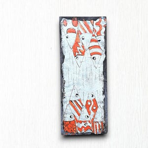 May include: A rectangular wooden plaque with a white background and a design of orange and white fish. The fish are arranged in a vertical pattern and have a stylised design. The plaque is signed "Elaine Rock" in the bottom left corner.