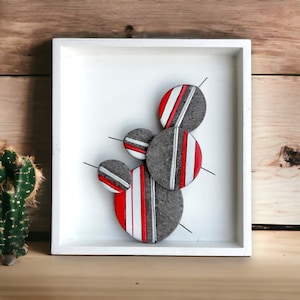 May include: A framed wall art piece featuring a cluster of five round, grey and white ceramic shapes with red stripes. The shapes are arranged in a vertical stack and are attached to the white background with thin metal rods.