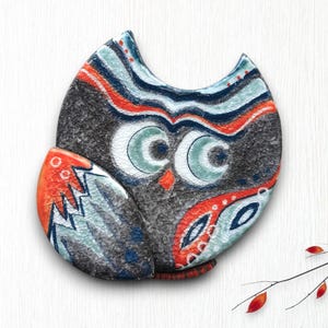 May include: A ceramic owl figurine with a textured, dark gray body and colorful accents. The owl features large, light blue eyes, orange and blue wings, and red and blue details. The design is a decorative, handcrafted item.