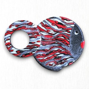 May include: A whimsical, hand-painted fish-shaped decorative item. The design features a circular ring attached to a rounded shape, both adorned with red, white, and gray wavy patterns. The fish has a gray body, a red lip, and a single eye.