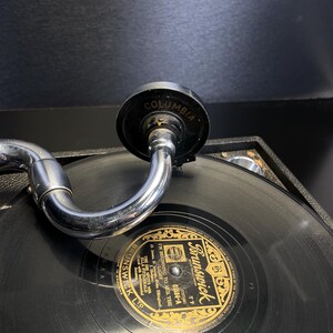 Puede incluir: Primer plano de un tocadiscos antiguo con un disco negro. La etiqueta del disco dice "Brunswick" en letras doradas. El brazo del tocadiscos es plateado y el cabezal es negro con "Columbia" impreso.