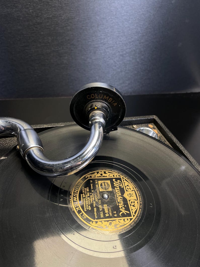Puede incluir: Primer plano de un tocadiscos antiguo con un disco negro. La etiqueta del disco dice "Brunswick" en letras doradas. El brazo del tocadiscos es plateado y el cabezal es negro con "Columbia" impreso.