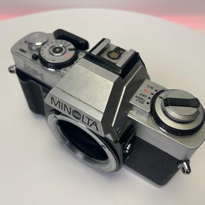 May include: A silver Minolta camera with black accents. The camera has a silver lens with a black ring around it. The camera has a black strap attached to it.