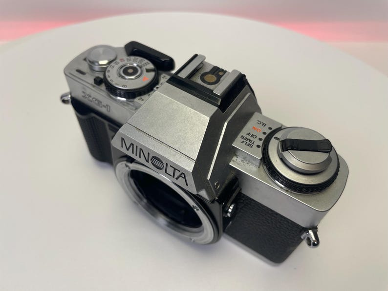 May include: A silver Minolta camera with black accents. The camera has a silver lens with a black ring around it. The camera has a black strap attached to it.