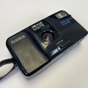 May include: Black Konica Pop Super auto flash focus free camera with a black strap. The camera has the Konica logo on the top left. The camera has a lens, flash, and an open slider.