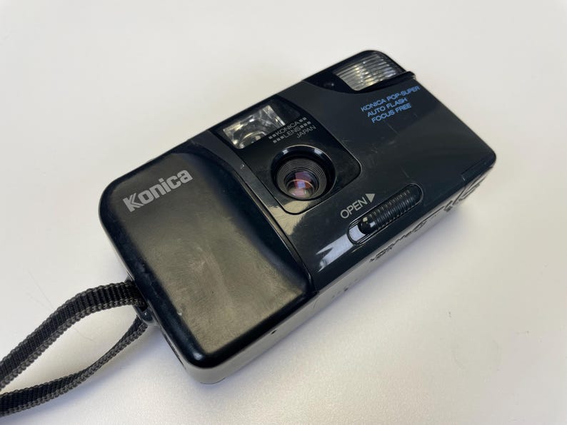 May include: Black Konica Pop Super auto flash focus free camera with a black strap. The camera has the Konica logo on the top left. The camera has a lens, flash, and an open slider.