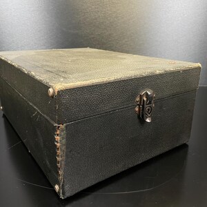 Puede incluir: Una caja negra rectangular con una superficie texturizada y un cierre met&aacute;lico. La caja tiene esquinas redondeadas y est&aacute; asegurada con peque&ntilde;os remaches met&aacute;licos. La caja parece ser vintage.