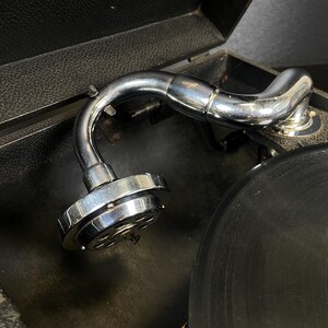 Puede incluir: Primer plano de un tocadiscos vintage con un brazo cromado brillante y un disco de vinilo negro. El tocadiscos est&aacute; alojado en una caja negra texturizada. El brazo es curvo y tiene un cartucho en el extremo.