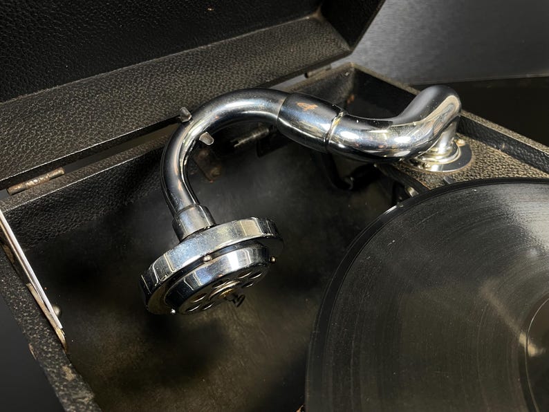 Puede incluir: Primer plano de un tocadiscos vintage con un brazo cromado brillante y un disco de vinilo negro. El tocadiscos est&aacute; alojado en una caja negra texturizada. El brazo es curvo y tiene un cartucho en el extremo.