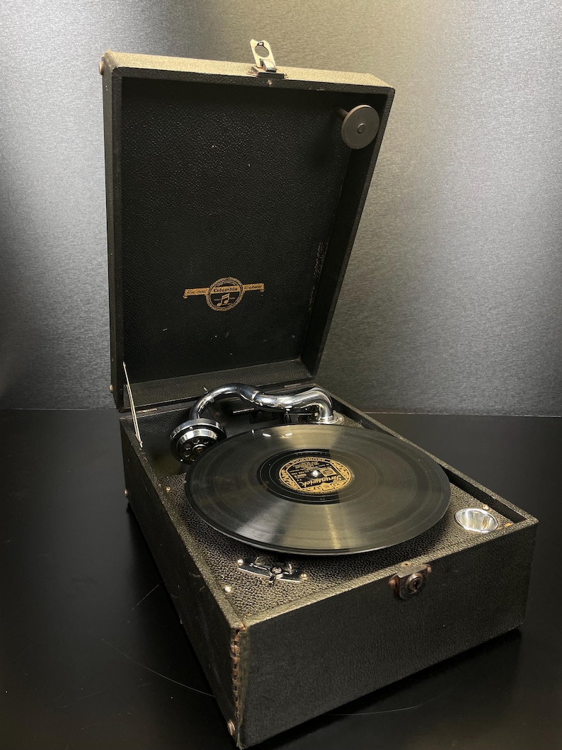 Puede incluir: Un tocadiscos port&aacute;til negro vintage con la tapa abierta, que revela un disco en el tocadiscos. El reproductor tiene un brazo plateado y un emblema dorado en el interior de la tapa. La caja es texturizada.