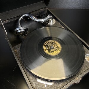 Puede incluir: Tocadiscos port&aacute;til negro vintage con brazo plateado y disco de vinilo. El tocadiscos est&aacute; en una caja negra con tapa abatible. La etiqueta del disco dice "Brunswick".