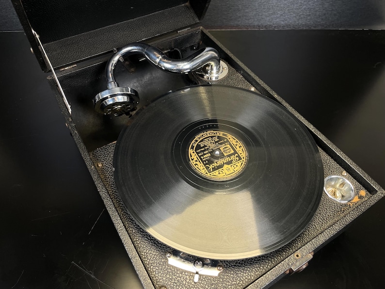 Puede incluir: Tocadiscos port&aacute;til negro vintage con brazo plateado y disco de vinilo. El tocadiscos est&aacute; en una caja negra con tapa abatible. La etiqueta del disco dice "Brunswick".
