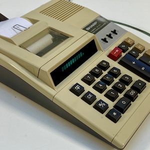 May include: A beige Sharp Compact 10-Digit Electronic Calculator with a built-in printer. The calculator has a large green digital display showing the number 8888888888. The calculator has a variety of keys including numbers, operators, and functions.