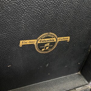 Puede incluir: Primer plano de una superficie negra texturizada con un emblema dorado. El emblema dice "Columbia Grafonola" con una nota musical y las palabras "Viva-tonal" y "Made in England".