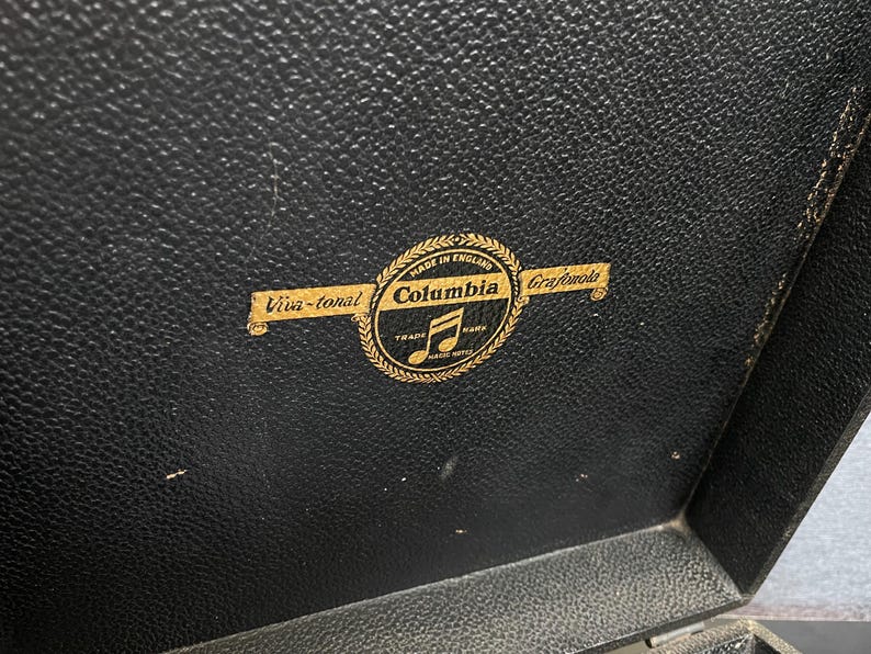 Puede incluir: Primer plano de una superficie negra texturizada con un emblema dorado. El emblema dice "Columbia Grafonola" con una nota musical y las palabras "Viva-tonal" y "Made in England".