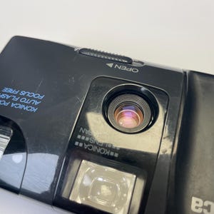 May include: Close-up of a black Konica Pop Super camera. The camera has a lens, flash, and the words "Konica Pop-Super Auto Flash Focus Free" printed in blue. The camera also has the word "Open" with an arrow.