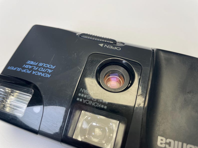 May include: Close-up of a black Konica Pop Super camera. The camera has a lens, flash, and the words "Konica Pop-Super Auto Flash Focus Free" printed in blue. The camera also has the word "Open" with an arrow.