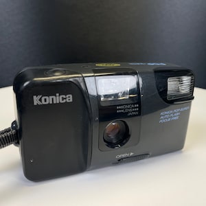 May include: Black Konica Pop Super 35mm film camera with a built-in flash. The camera has the brand name "Konica" printed on the front. The camera has a lens and the words "AUTO FLASH FOCUS FREE" are printed on the side.